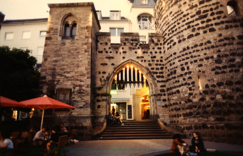 A stone medieval city gate with an arched entrance stands among modern buildings. People sit at outdoor tables under red umbrellas and on the ground, enjoying the evening. Warm lights glow from shops inside the gate.
