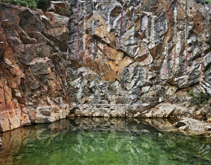 A rocky, vertical quarry wall with jagged textures and reddish-brown hues rises above a pool of green water, with reflections of the rock face visible on the water’s surface. Sparse greenery is seen at the edges.