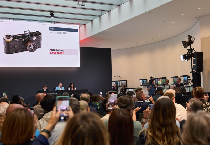 A large crowd watches an auction in a modern room as a vintage Leica camera is displayed on a screen with a current bid of 6,000,000 euros. Many audience members are raising phones to take photos.