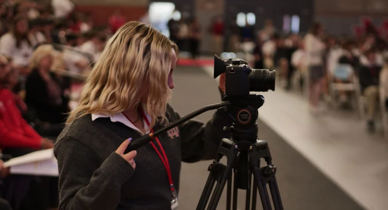 A person with wavy blonde hair operates a video camera on a tripod at an indoor event, facing a crowd seated in the background. The individual wears a gray sweater and a red lanyard.