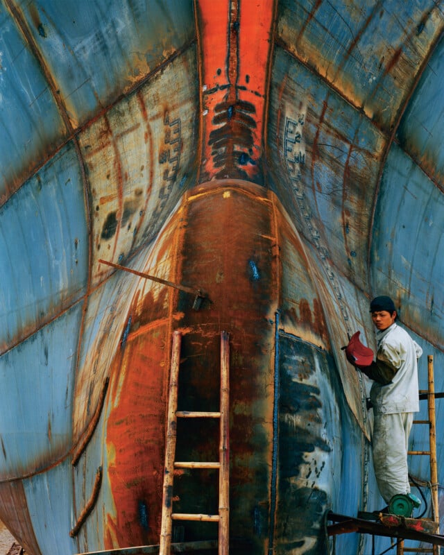 A worker in white overalls stands on a ladder, sanding or painting the rusted, blue and orange hull of a large ship in dry dock. The ship towers above, showing its weathered surface and bold colors.