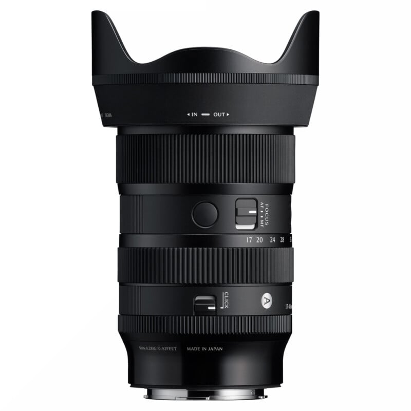 A black Sigma camera lens with zoom and focus rings, lens hood attached, and autofocus/manual switch visible. The lens displays focal lengths and various markings, standing upright against a white background.