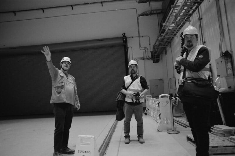 Three men in safety gear and hard hats stand in an industrial setting. One man raises his hand, another holds a camera, and a warning sign labeled "Cuidado" is in the foreground. The scene is in black and white.