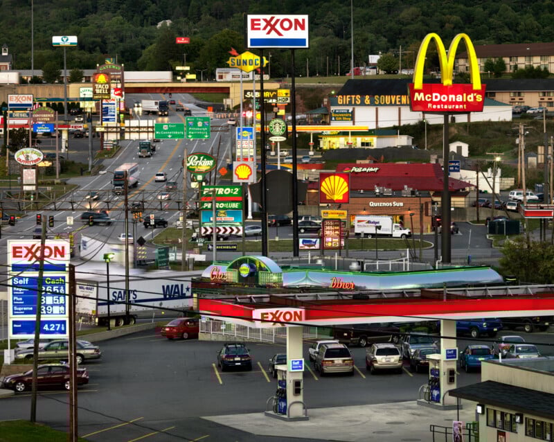 A busy roadside scene features numerous gas stations, fast food restaurants, and bright signs, including Exxon, McDonald’s, Shell, and Sunoco, with parked cars and traffic on intersecting streets.