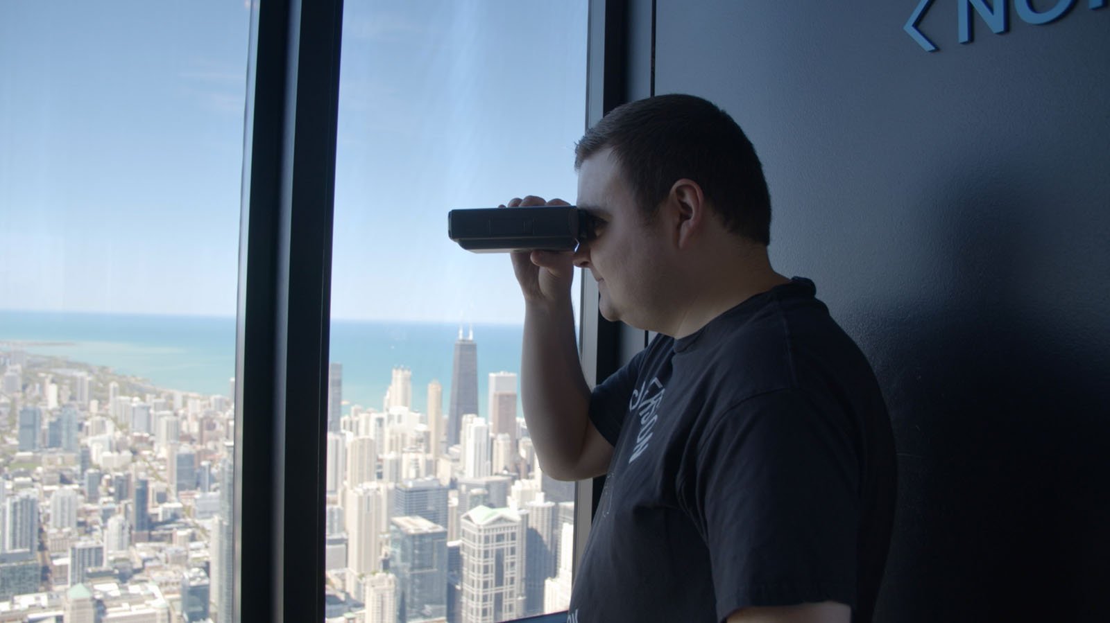 A person looks through a coin-operated binocular viewer inside a tall building, overlooking a cityscape with skyscrapers near a large body of water under a clear sky.