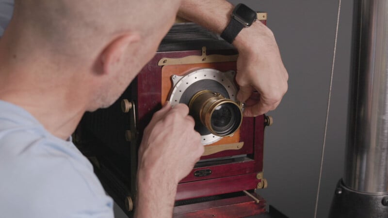 A person adjusts the brass lens of a vintage large format camera, seen from behind. The camera has a wooden body and metal fittings, and the person is wearing a smartwatch on their left wrist.