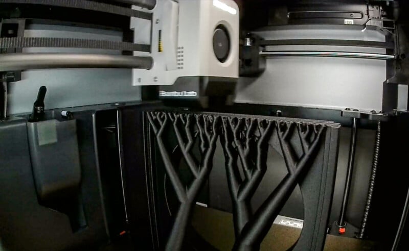 A 3D printer is in action, printing a complex object with multiple branching, black vertical supports inside the printer’s enclosed chamber. The printer head is visible near the top.
