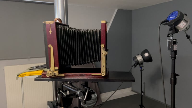 A large format wooden bellows camera is mounted on a stand in a photography studio, surrounded by studio lights and equipment against gray walls.