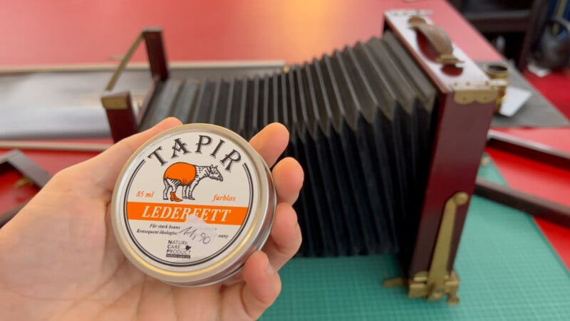 A hand holds a tin of Tapir Lederfett leather care product in front of a vintage large format camera with a bellows, on a table with a green cutting mat and red surface.
