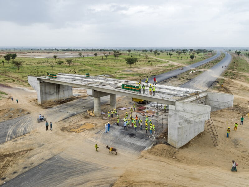 A group of construction workers wearing yellow vests work on an unfinished concrete bridge over a dirt road in a rural area, with open fields and a paved road extending into the distance.