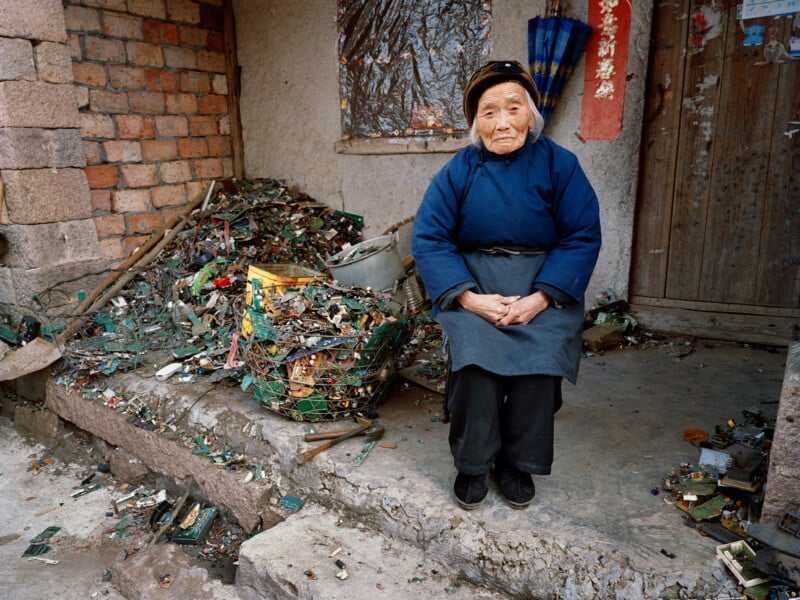 An elderly woman in a blue coat and black hat sits on a worn stone step outside a brick house, surrounded by piles of discarded electronic circuit boards and debris.