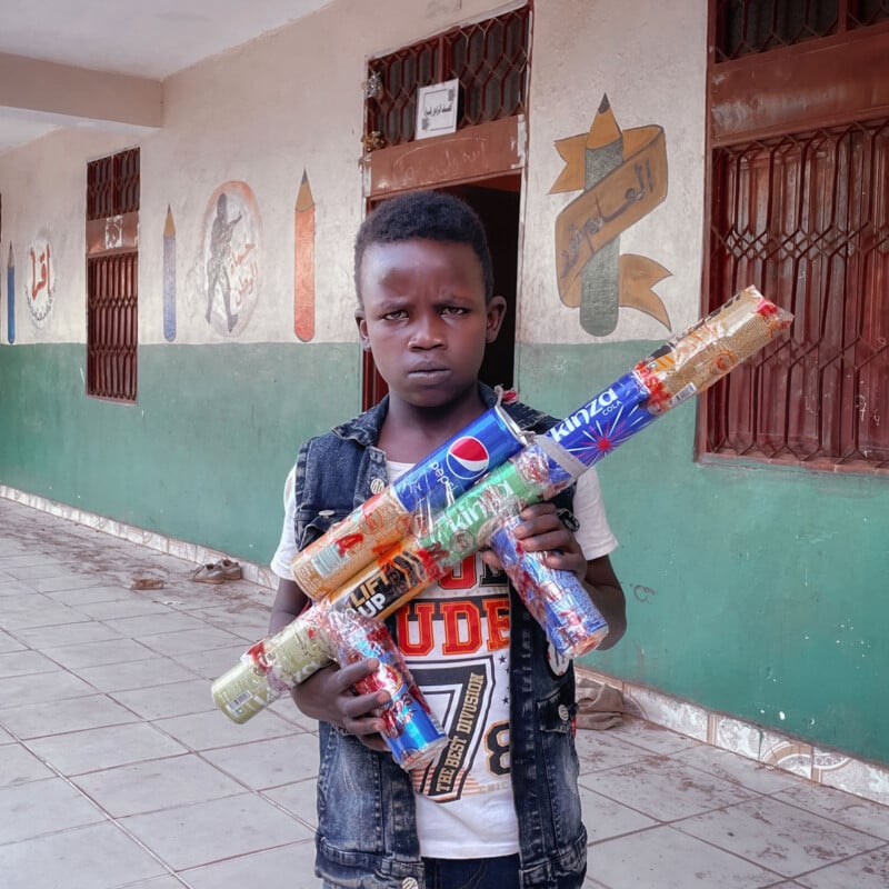 A young boy stands outside a building, holding a large toy gun made from colorful recycled cans. The wall behind him is decorated with painted art and Arabic script. The boy looks directly at the camera with a serious expression.