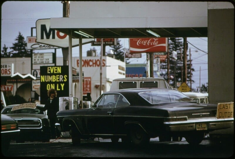 A woman holds a sign reading "EVEN NUMBERS ONLY" at a crowded 1970s gas station, with vintage cars lined up and various signs and advertisements visible in the background.