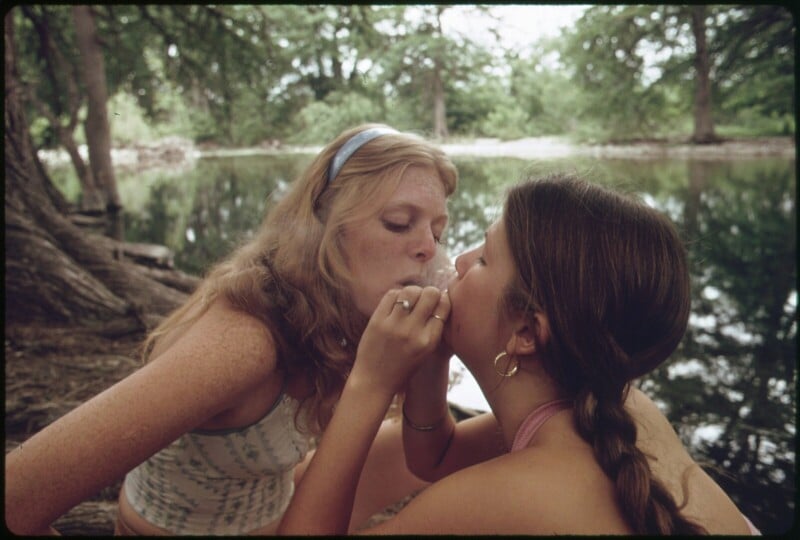 Two young women sit by a riverside; one holds a small object to the other's lips as they both concentrate. Trees and calm water are in the background, creating a peaceful outdoor setting.