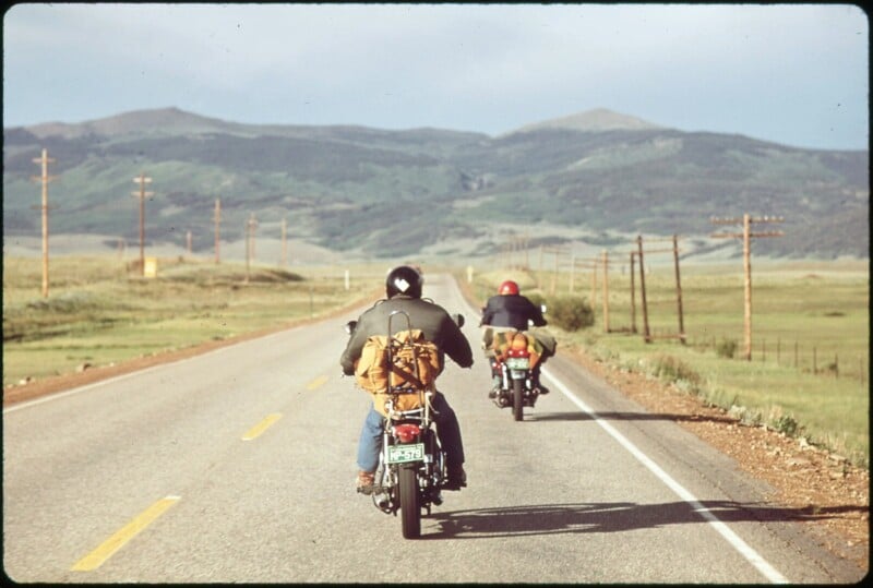Two people on motorcycles ride down an empty highway through open countryside, with mountains in the distance and power lines running alongside the road.