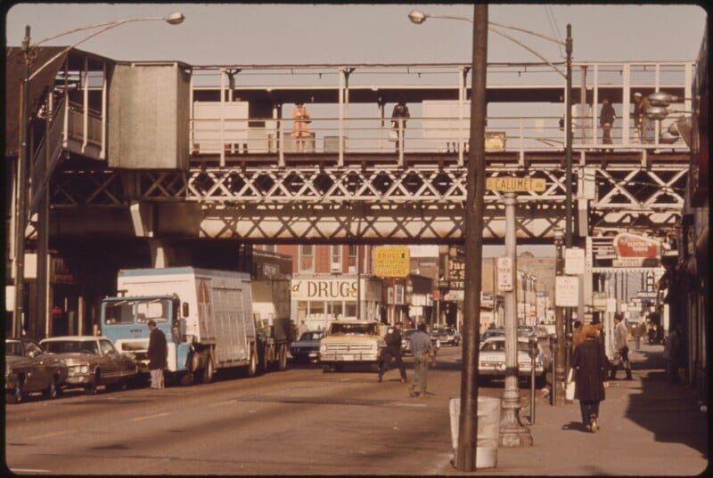 A busy urban street with cars, trucks, and pedestrians, featuring an elevated train track overhead and people walking on the sidewalk; storefronts and street signs are visible in the background.