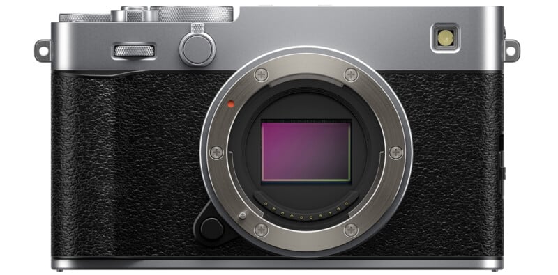 A digital mirrorless camera without a lens, showing the sensor and metal lens mount. The camera body is silver and black with a textured grip and various control dials and buttons.