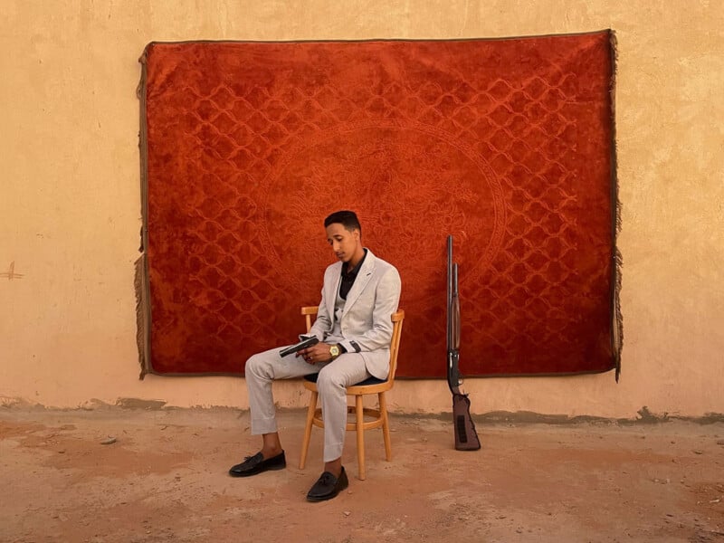 A man in a light gray suit sits on a wooden chair against a red patterned rug hung on a beige wall. He reads a book, and a long rifle leans upright against the chair beside him.