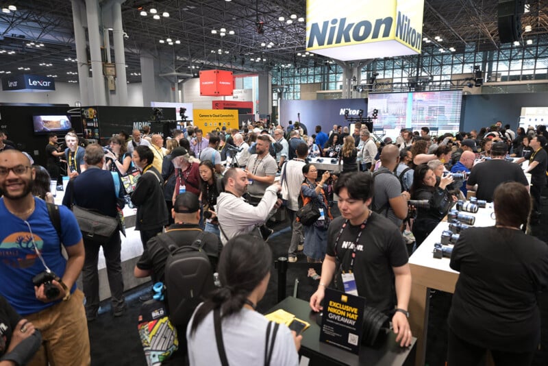 A large crowd gathers at a busy Nikon booth during a technology convention, with people testing cameras, taking photos, and interacting with booth staff under prominent Nikon signage.