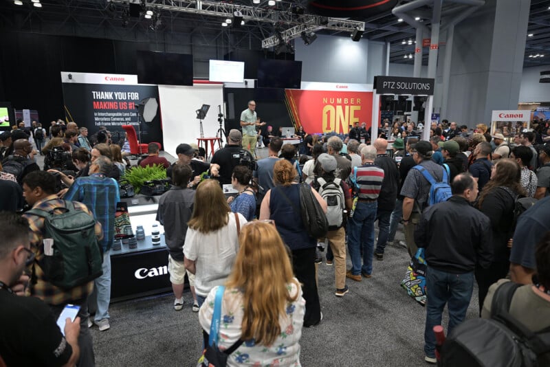 A large crowd gathers around a Canon booth at a convention, listening to a speaker on stage. People wear backpacks and cameras, and banners and equipment displays are visible around the busy event space.