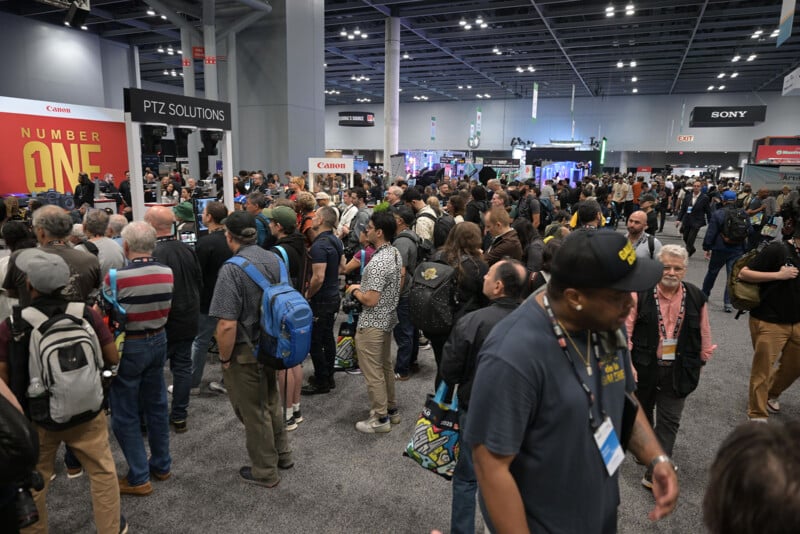 A large crowd of people, many with backpacks and cameras, gathers at an indoor technology or photography expo. Booths with company logos like Canon and Sony are visible in the background.