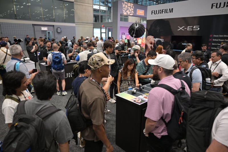 A large group of people gathers around a display booth for the FUJIFILM X-E5 camera at a convention. Many attendees carry backpacks and cameras, while some interact with products on the counter.