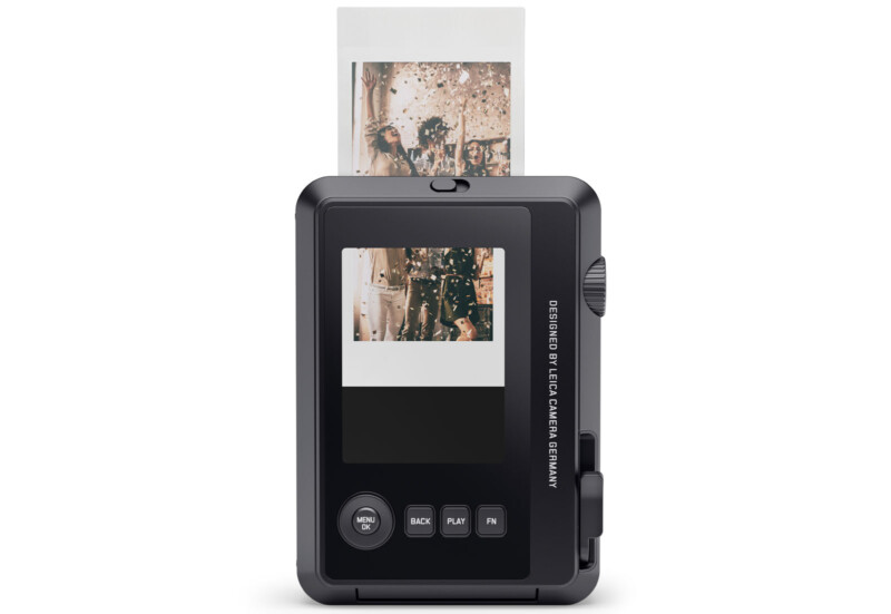 A digital instant camera prints a photo from its top slot. The camera has a display screen showing the image, along with menu, back, play, and print buttons below the screen.