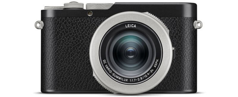 Front view of a compact black Leica digital camera with a textured grip and a silver-rimmed lens. The lens features branding and technical details, and the camera body has a minimalist, classic design.