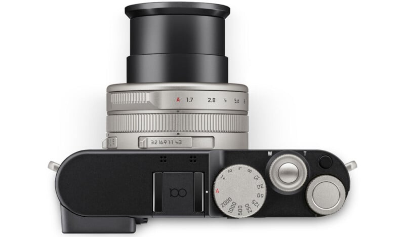 Top view of a silver and black digital camera with a lens extended, showing various dials and buttons for settings like aperture, shutter speed, and zoom.