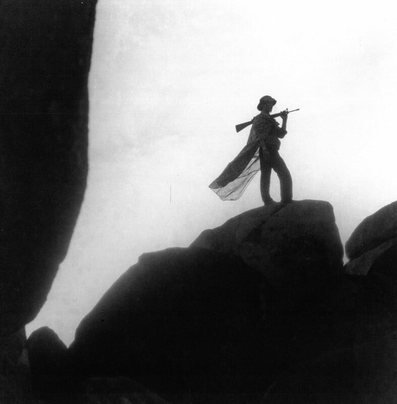 Silhouette of a person standing on large rocks, holding a rifle and wearing a flowing cape, set against a bright, cloudy sky.