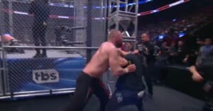 A shirtless wrestler pushes a man in dark clothing against a cage beside a wrestling ring during a live event, with a crowd watching in the background. The TBS logo is visible on the cage.