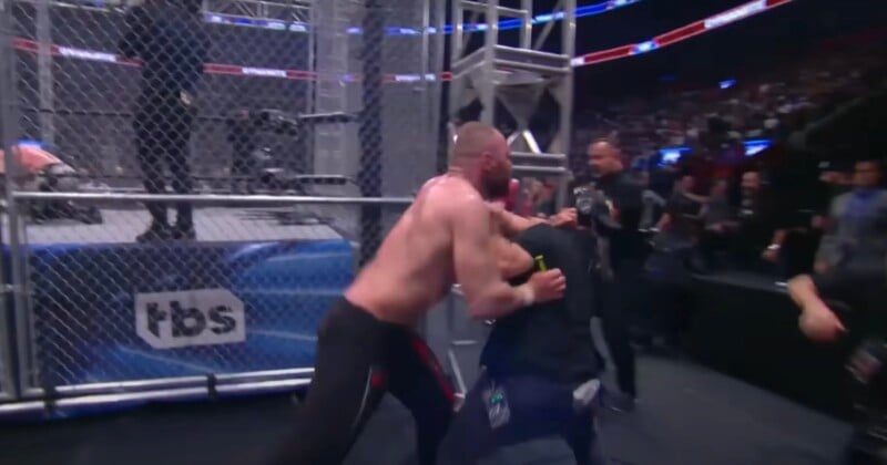 A shirtless wrestler pushes a man in dark clothing against a cage beside a wrestling ring during a live event, with a crowd watching in the background. The TBS logo is visible on the cage.