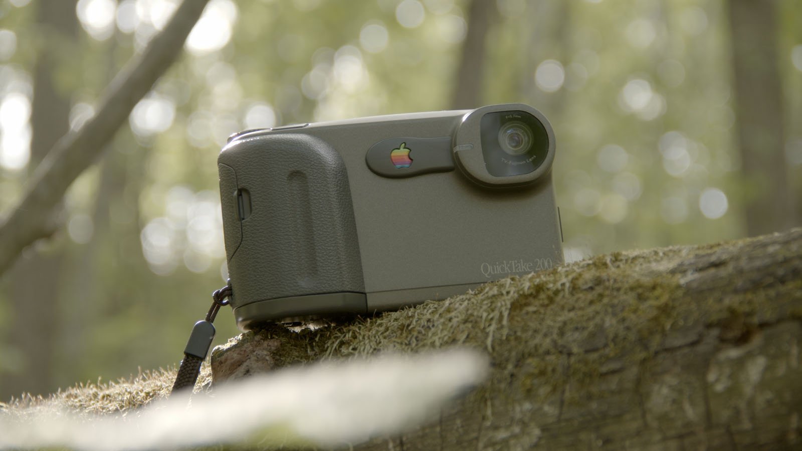 A vintage Apple QuickTake 200 digital camera rests on a mossy tree trunk in a sunlit forest setting.