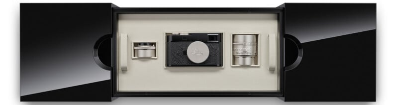 A luxury box set displaying a black Leica camera with a lens cap in the center, flanked by two silver camera lenses and accessories, all set in a cream-colored interior with black exterior doors partially open.