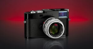 A black Leica camera with a textured grip and silver lens is displayed on a reflective surface against a gradient red and black background.