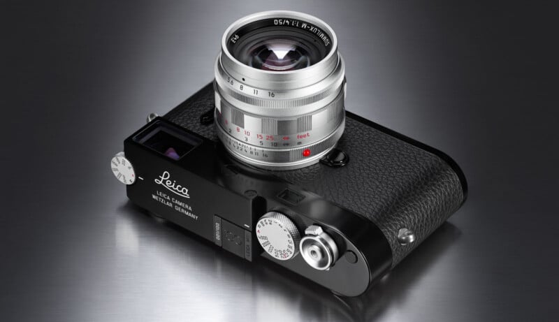 A silver and black Leica camera with a textured grip and prominent lens is placed on a reflective metallic surface, viewed from an angle showing the top and front of the camera.