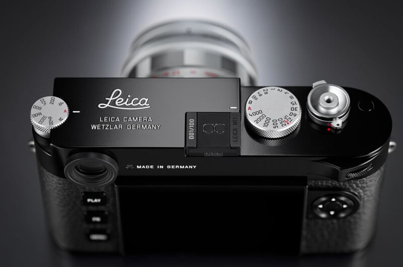 Top view of a black Leica camera, showing the engraved Leica logo, control dials, and buttons. The lens is partially visible, and the camera is placed on a reflective dark surface.