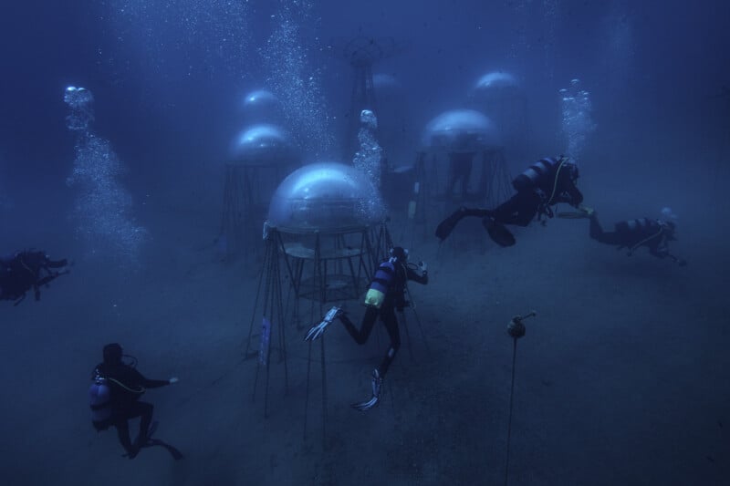Several scuba divers swim around dome-shaped underwater structures on the ocean floor, with bubbles rising to the surface in the deep blue water. The scene suggests an underwater research or habitat site.