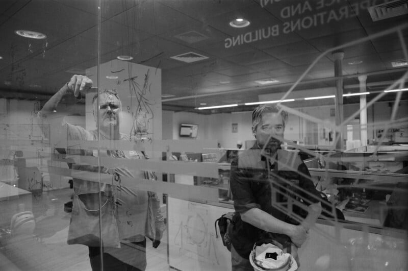 Two men stand behind a glass wall with diagrams and reflections visible; one is drawing or writing on the glass, while the other stands with his arms crossed, holding a hard hat. An office environment is seen in the background.