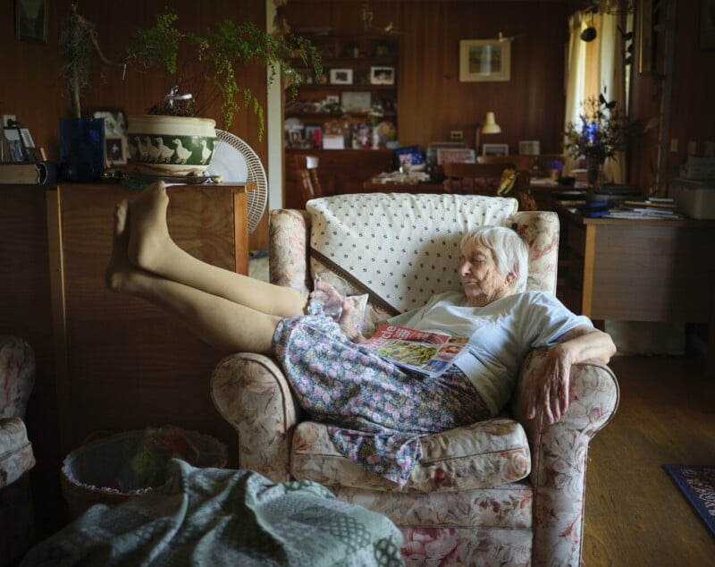 An elderly woman rests in a patterned armchair with her legs up, eyes closed, and a magazine on her lap. The cozy living room around her is filled with personal items, plants, and warm natural light.