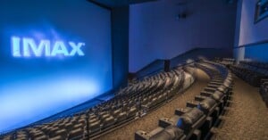 A modern IMAX theater with a large, curved screen displaying the IMAX logo, surrounded by rows of empty, plush black seats with cup holders. The room is dimly lit, creating a cinematic atmosphere.