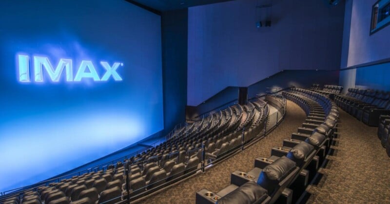 A modern IMAX theater with a large, curved screen displaying the IMAX logo, surrounded by rows of empty, plush black seats with cup holders. The room is dimly lit, creating a cinematic atmosphere.
