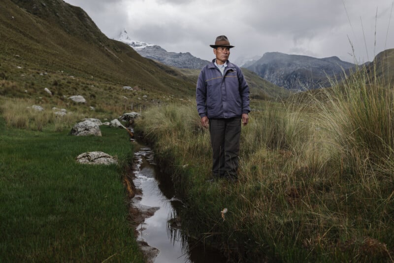 A person in a hat and jacket stands beside a narrow stream in a grassy mountain valley, with cloudy skies and distant snow-capped peaks in the background.