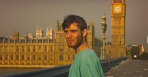 A man in a hospital gown stands on an empty bridge in front of the Houses of Parliament and Big Ben in London, with scattered debris on the ground and a hazy sky.