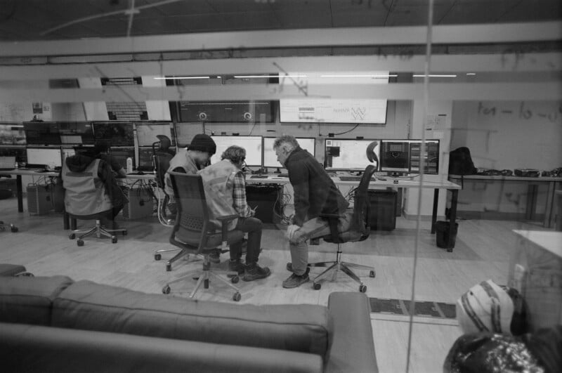 Black and white photo of four people working at computer desks in a modern office, with three seated together discussing something, seen through a glass wall with writing on it. A sofa is in the foreground.