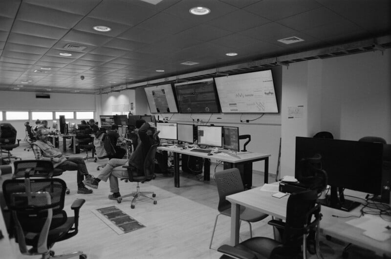 A modern office with people sitting at desks and monitors, large screens displaying data on the wall, and empty chairs around. The room has a high ceiling with fluorescent lighting and a minimalist design.