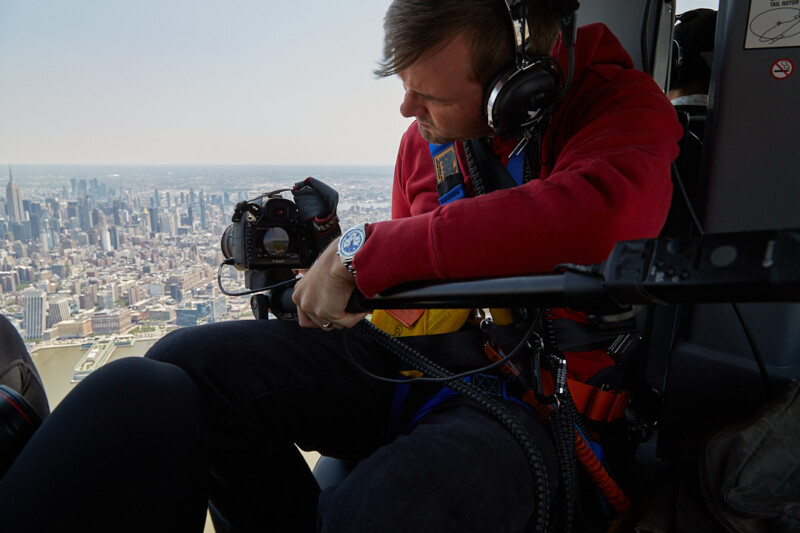 A man wearing headphones and a red hoodie operates a camera while seated inside a helicopter, looking out over a city skyline with skyscrapers visible in the background.