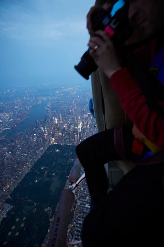 A person holding a camera leans out of a helicopter above New York City at dusk, capturing a view of Central Park and the cityscape far below.