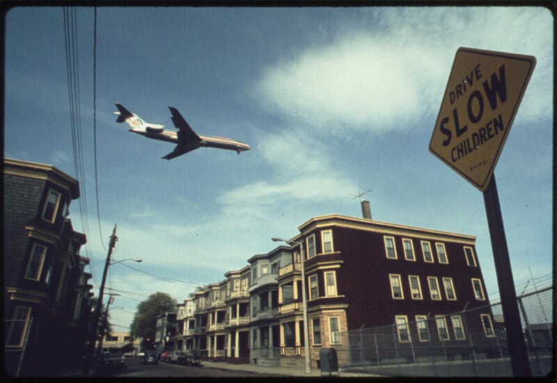 A low-flying commercial airplane passes over a residential neighborhood with multi-story houses; a yellow "Drive Slow Children" sign is visible in the foreground under a blue sky with scattered clouds.