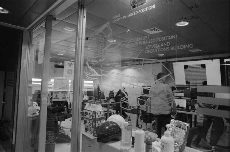 Black and white photo of an office with people working at computers, safety helmets and sanitizer bottles on a table, and large diagrams and safety signs visible on the glass wall in the foreground.